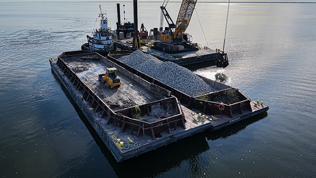 A close view of an empty barge with a front-end loader sitting idle. A tug pushing a barge loaded with cobble is visible, along with a third barge supporting a large crane. The tug and barges are surrounded by calm water that reflects the sunshine.